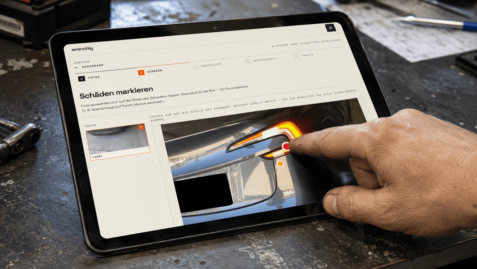 Annehmer markiert mit dem Finger einen Schaden direkt auf dem Foto im wrenchly-Annahmeprotokoll — Tablet auf einer Werkbank in der Kfz-Werkstatt